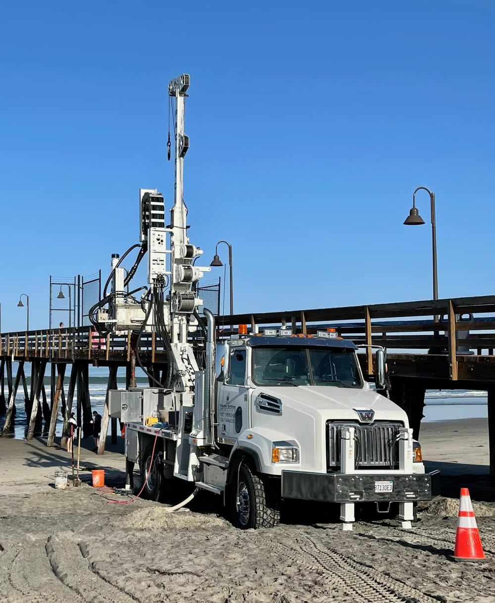 A Pacific Drilling truck rig with its mast raised on the sand beside a pier, crew members working at the shoreline