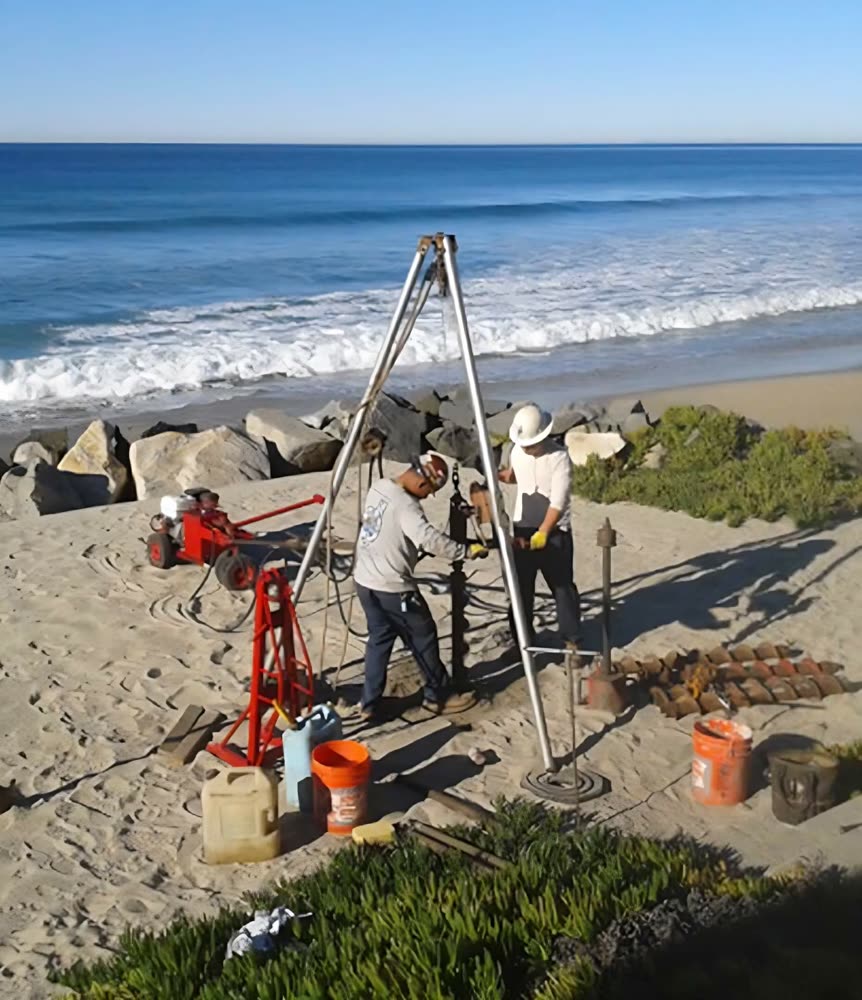A tripod rig set up on a beach bluff, two crew members working alongside it